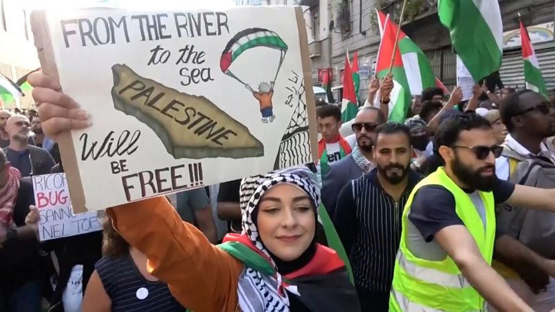 An anti-Israel protestor holds a sign depicting a Hamas paraglider and the slogan "From the River to the Sea", which calls for the death of all Israelis.
